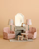 The Maileg Living Room Set includes two pink plush armchairs, a wooden table, mouse dollhouse furniture, a grey sideboard with mirror, two floral lamps, an alarm clock, and a tiny dog figurine—all set against a beige background.