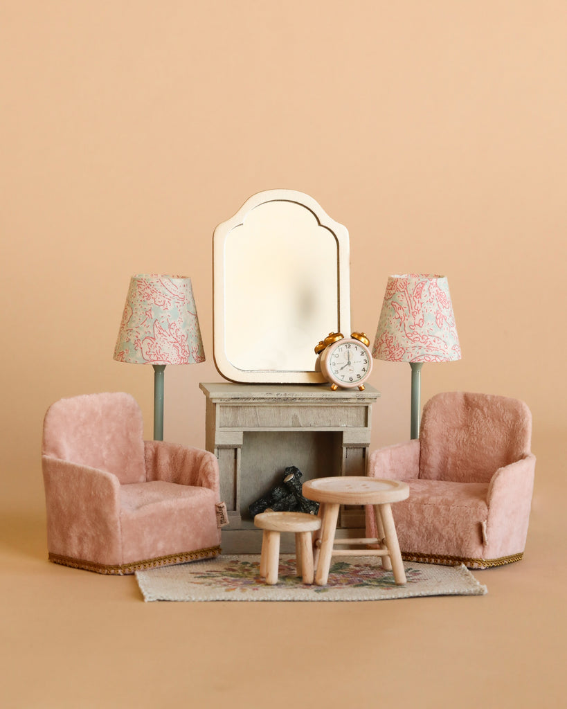 The Maileg Living Room Set includes two pink plush armchairs, a wooden table, mouse dollhouse furniture, a grey sideboard with mirror, two floral lamps, an alarm clock, and a tiny dog figurine—all set against a beige background.