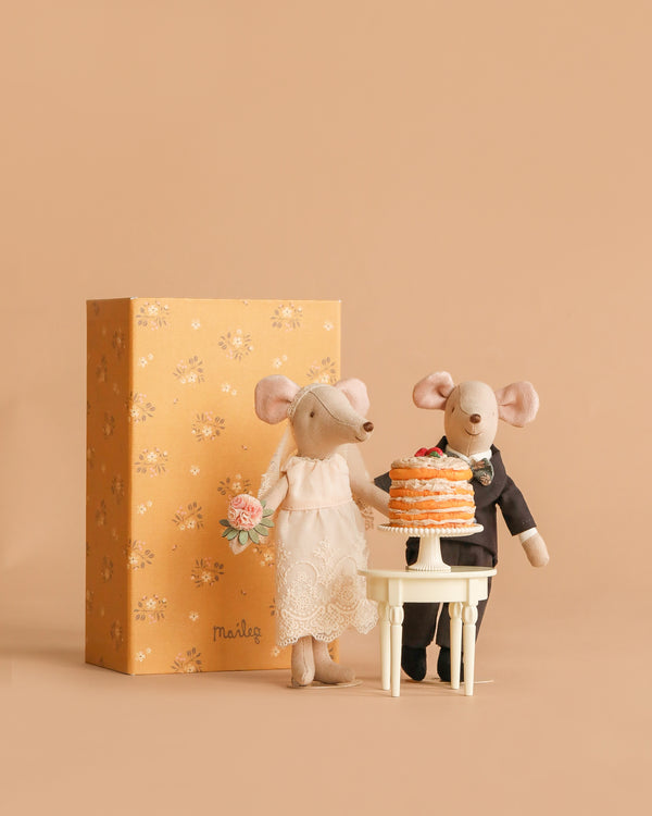 The Maileg Wedding Day Set features two mice dressed as bride and groom, a mini side table with a party cake, and an orange floral box for display. The bride holds a bouquet in lace while the groom wears a suit.