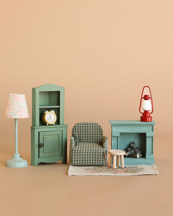 The Maileg Living Room Set features a plaid armchair, teal lamp, green cabinet with clock, Maileg table and stool, rug, and mouse fireplace with logs and lantern—all arranged against a beige background.