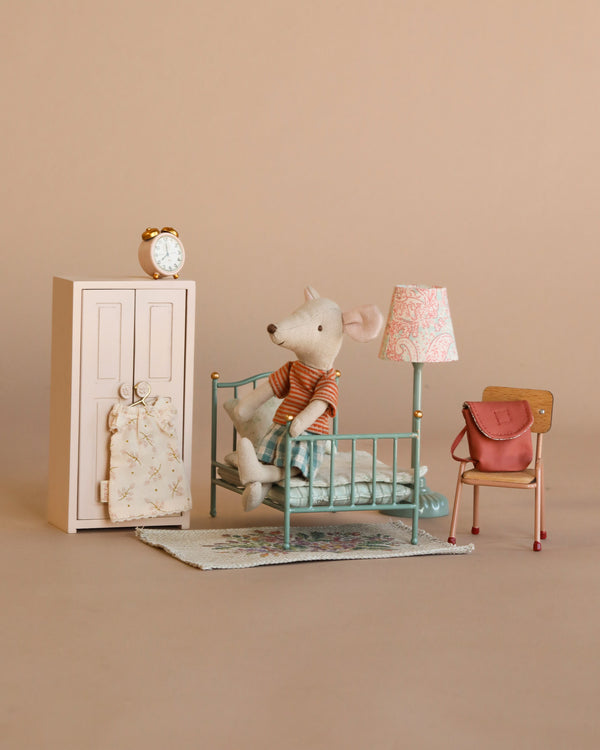 The Maileg Little Sister Bedroom Set features a small mouse doll in a nightgown on a mint green bed with blue quilt, alongside accessories like a pink wardrobe, vintage lamp, chair with red bag, and clock on a beige background.