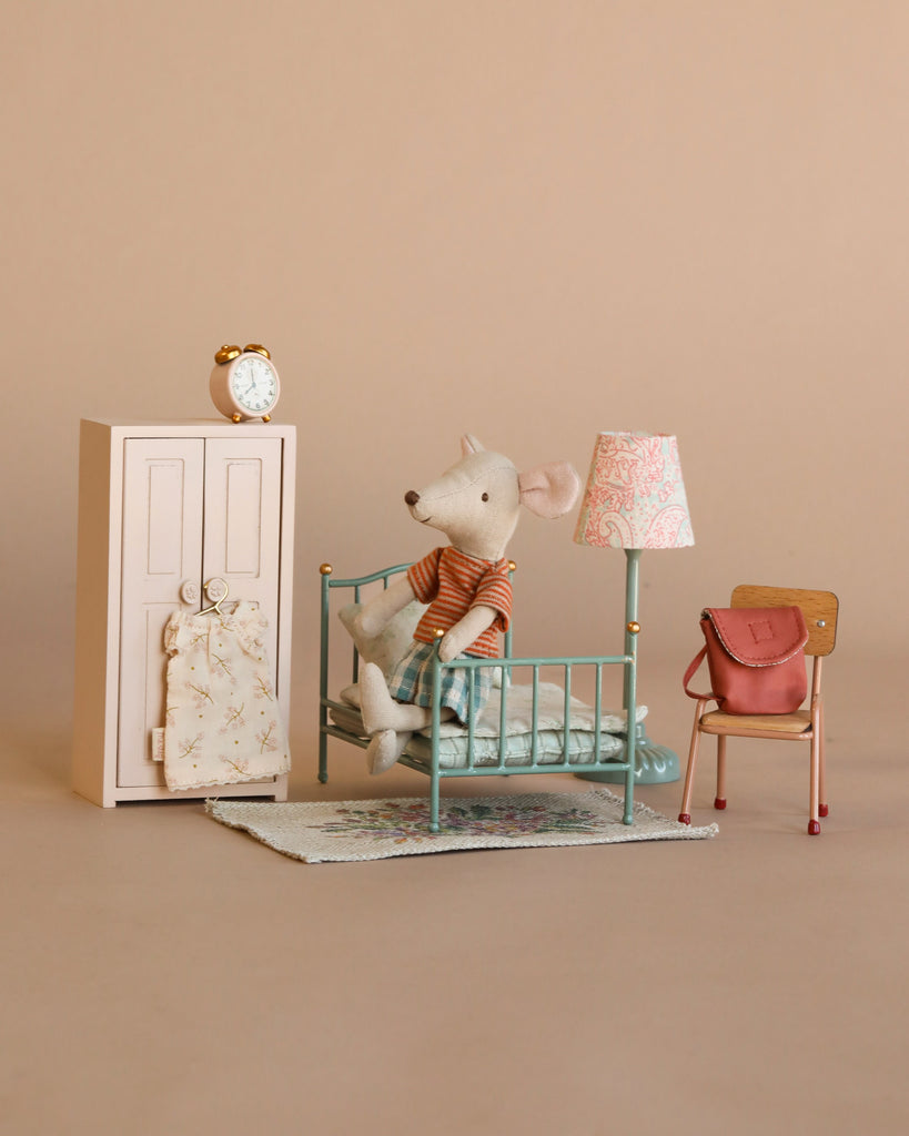 The Maileg Little Sister Bedroom Set features a small mouse doll in a nightgown on a mint green bed with blue quilt, alongside accessories like a pink wardrobe, vintage lamp, chair with red bag, and clock on a beige background.