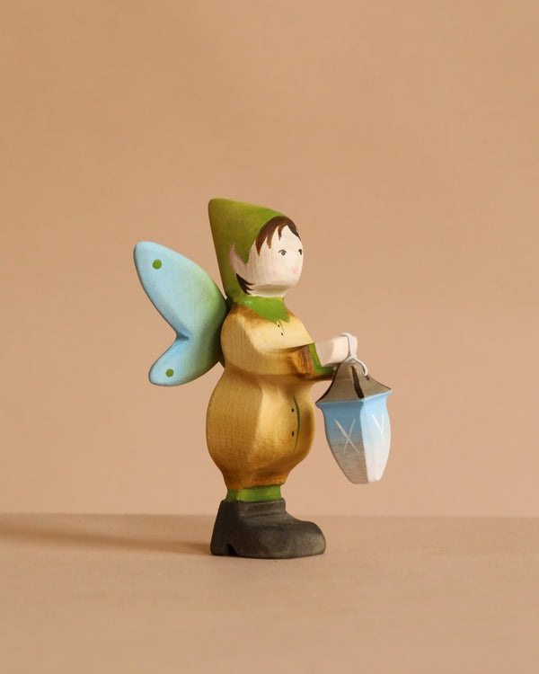The Bumbu Handmade Wooden Winged Elf with Lamp Set features a small, handcrafted wooden elf fairy with blue wings, a green hat, tan clothes, and a blue-and-white lantern set against a plain beige background.