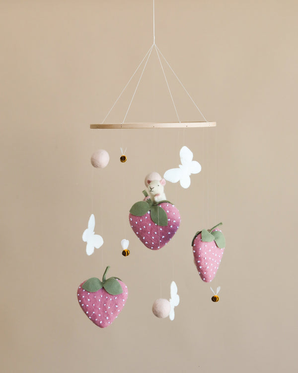 A nursery mobile featuring plush strawberries, white butterflies, and felt balls hanging from a wooden hoop against a beige background. A small figurine rests on one of the strawberries. Handmade Mobile - Field of Strawberries - Final Sale.