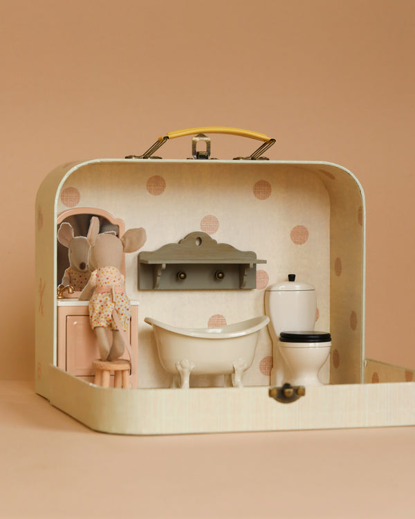 The Maileg Bathroom Starter Set is neatly presented inside an open suitcase and features a mouse doll dressed in a floral dress. The set includes a bathtub, toilet, and suitcase sink with cabinets, all against a light peach background with dotted patterns decorating the walls of the suitcase.
