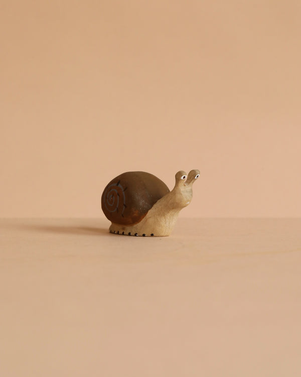 The Bumbu Handmade Wooden Snail, a small brown wooden figurine with a spiral shell, sits on a beige surface against a matching background, evoking the charm of handcrafted design.