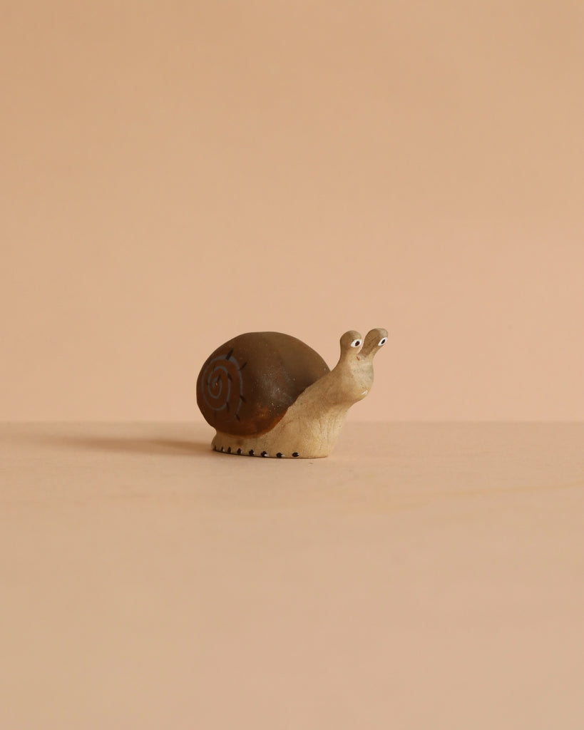 The Bumbu Handmade Wooden Snail, a small brown wooden figurine with a spiral shell, sits on a beige surface against a matching background, evoking the charm of handcrafted design.