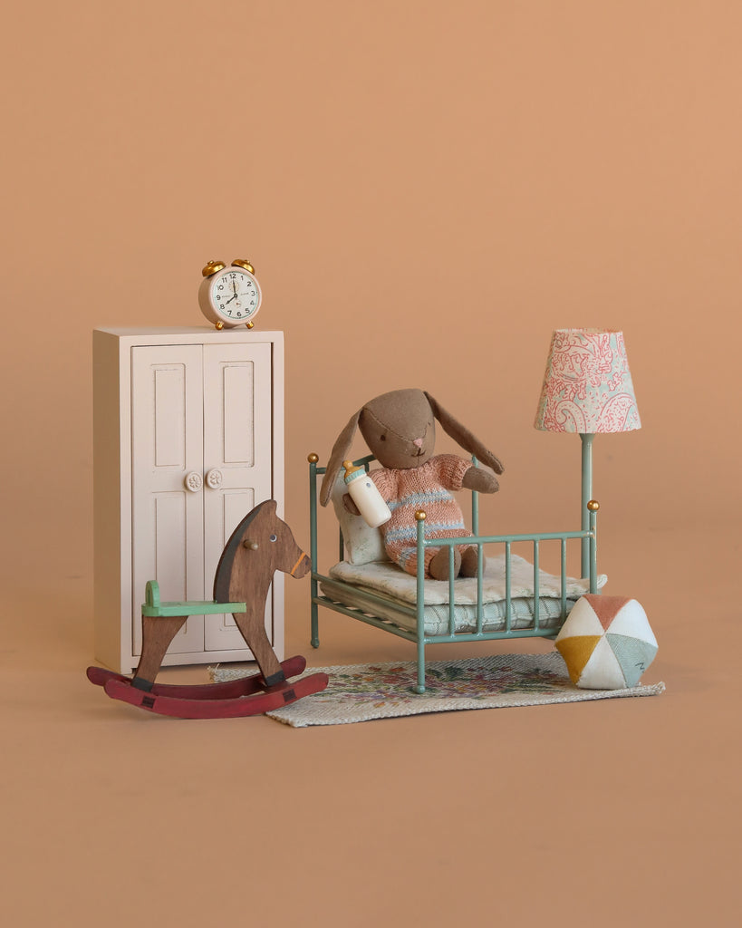 The Maileg Micro Bunny Bedroom Set features a miniature bedroom with a Maileg Bunny in a vintage bed, holding a bottle, plus tiny accessories like an alarm clock, rocking horse, rug, and lamp—all on a peach background.