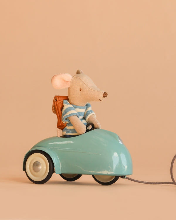 A Maileg Big Brother Adventure Set mouse, dressed in a striped shirt and backpack, sits in a small blue Maileg Mouse Car with white wheels against a plain beige background.