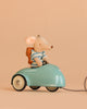 A Maileg Big Brother Adventure Set mouse, dressed in a striped shirt and backpack, sits in a small blue Maileg Mouse Car with white wheels against a plain beige background.