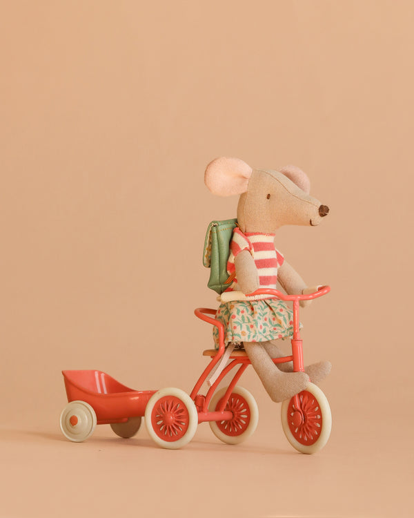 The Maileg Big Sister Tricycle Set features a mouse in a striped shirt, floral skirt, and backpack, riding a red tricycle with a matching wagon against a plain beige background.