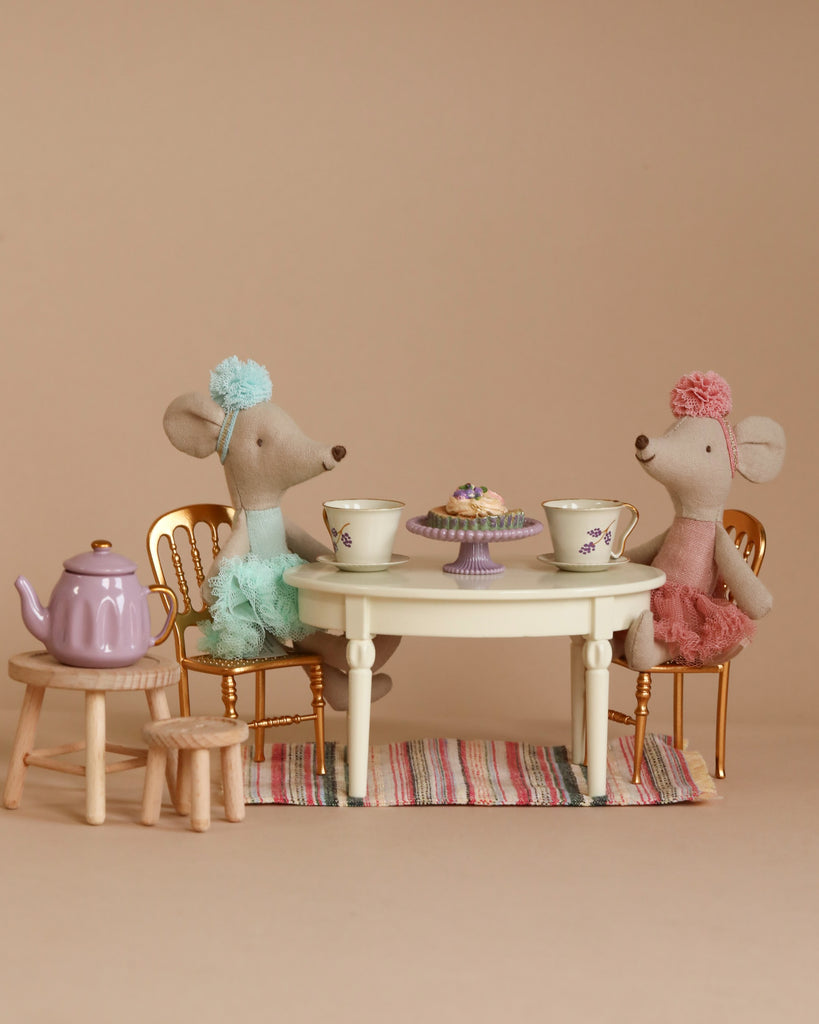 Two Maileg ballerina mice, elegantly dressed in pastel outfits with matching headpieces, are seated on gold chairs at a small round table. Each mouse has a teacup from the Maileg Tea Party Starter Set in front of them, while a charming purple teapot sits on a nearby wooden stool and an assortment of pastries is displayed on a stand in the center of the table.
