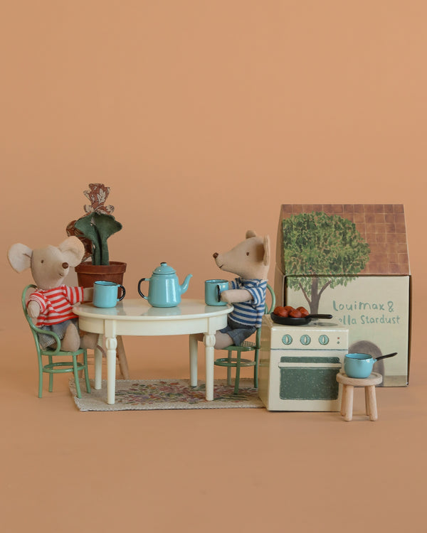 The Maileg LouiMax and Stella Cozy Kitchen Set features two toy mice at a table with blue dishes and a teapot, plus a toy stove and potted plant, creating a cozy Scandinavian kitchen scene against a light brown background.