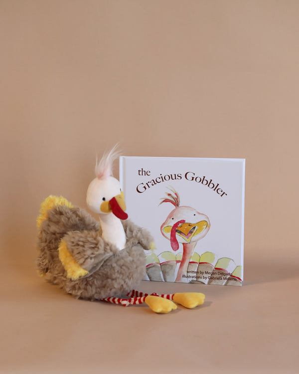 A plush turkey toy beside a children's book titled "The Gracious Gobbler Bundle: Children's Book, Plush and Cards" on a neutral background, embodying a Thanksgiving tradition. The turkey has a white head, grey body, and