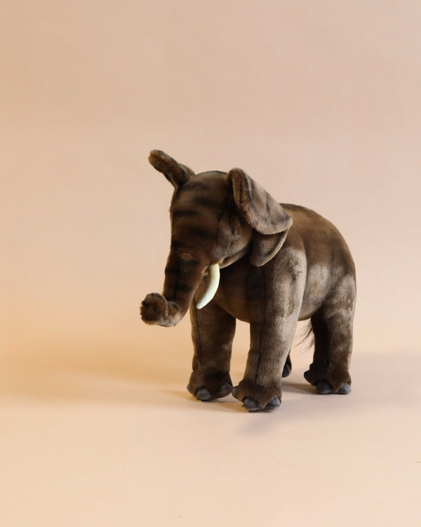 A brown, plush Elephant Stuffed Animal with large ears, tusks, and a raised trunk stands on a neutral beige background. The hand-sewn animal appears soft and huggable, with detailed stitching and a realistic design, making it a unique handcrafted toy.