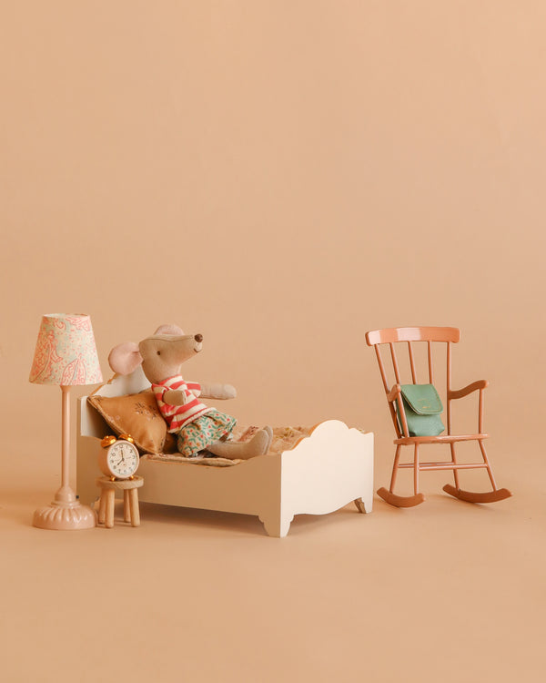 The Maileg Big Sister Bedroom Set features a plush mouse in striped pajamas on a white bed, with accessories like a patterned lamp, vintage-style clock, pink rocking chair, and green bag for a charming dollhouse scene.