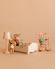 The Maileg Big Sister Bedroom Set features a plush mouse in striped pajamas on a white bed, with accessories like a patterned lamp, vintage-style clock, pink rocking chair, and green bag for a charming dollhouse scene.