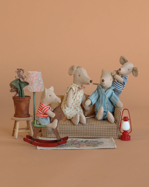 Four Maileg mice, dressed in colorful clothes, relax with the Maileg Family Room Set—featuring a couch, rocking horse, potted plant, lamp, rug, and lantern—arranged against a plain light brown background.