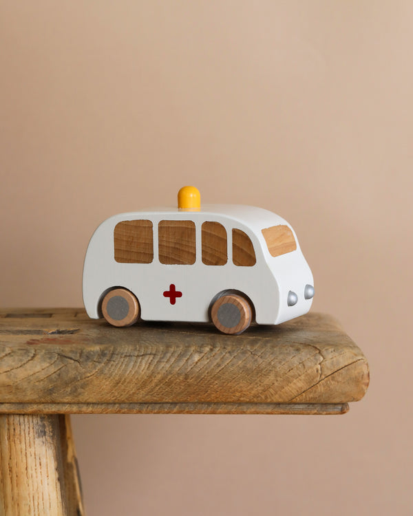 The Maileg Wooden Ambulance, crafted from solid wood, features a white body, brown windows, gray wheels, and an orange top light. It includes a red cross on the side and sits charmingly on a rustic wooden surface against a light brown background.