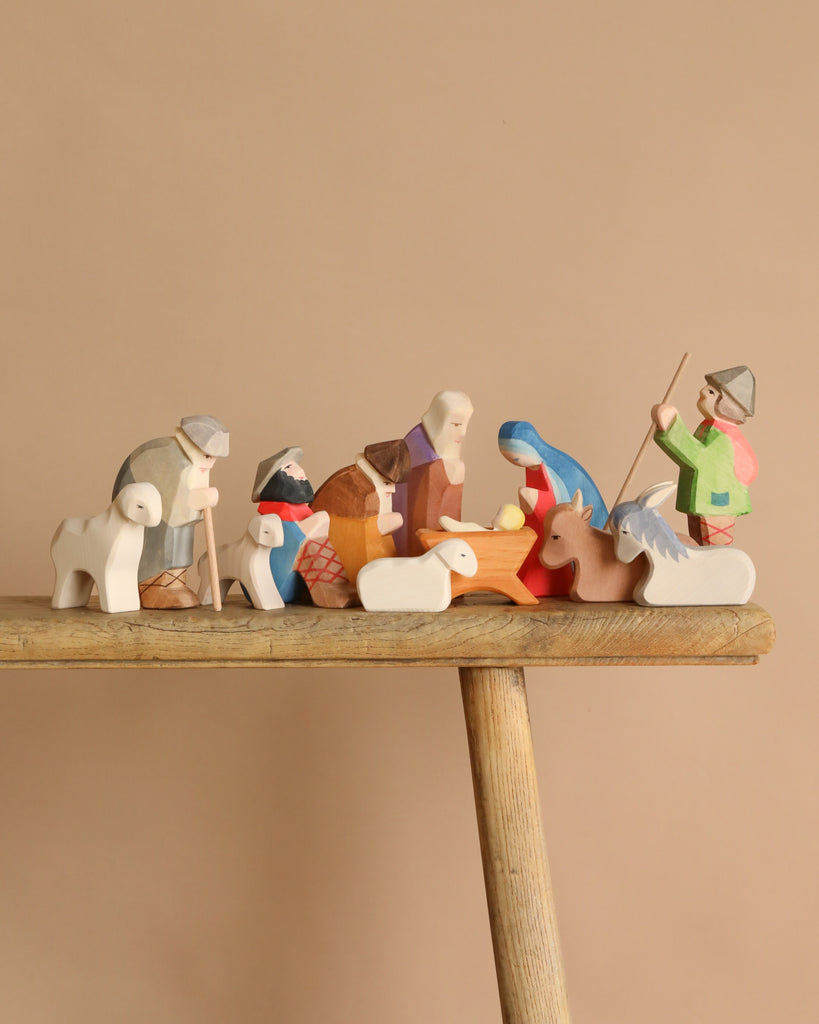 The Ostheimer Nativity Set - 13 Pieces features hand-painted wooden figures like Mary, Joseph, baby Jesus, shepherds, animals, and wise men on a rustic stool. Hand-crafted in Germany in the Waldorf tradition against a beige background.