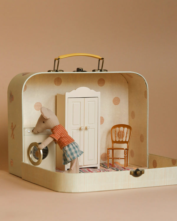 The Maileg Laundry Room Starter Set features a charming miniature scene where a mouse doll, dressed in a plaid skirt and striped shirt, is doing laundry in front of a washing machine. The room includes a white wardrobe, rug, and golden chair, all delightfully arranged within an endearing Maileg suitcase.