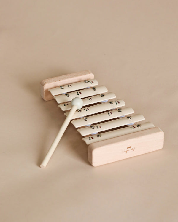 A small Cherry wooden xylophone with white metal bars and a beech wood frame, accompanied by a single mallet, positioned on a pale beige background.