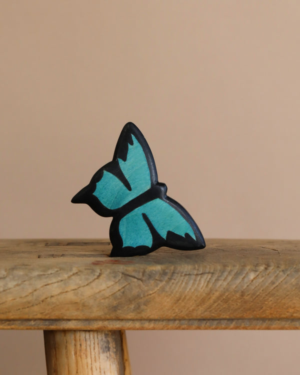 The Handmade Holzwald Butterfly, a small, blue and black painted wooden decoration, rests on a rustic surface against a beige background, embodying the charm of sustainable toys.