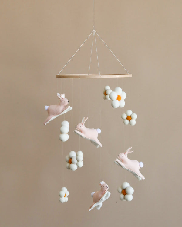 A handmade nursery mobile featuring three Jumping Bunnies and clusters of white and soft yellow pompoms, hanging against a light brown background.