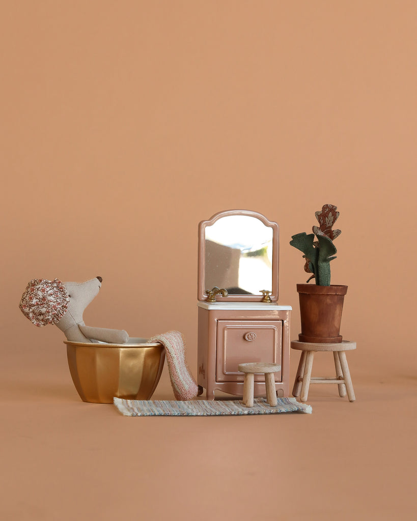 A Maileg Cozy Bathroom Set features a wellness mouse relaxing in a golden bathtub, a pink sink dresser with mirror, a stool, flower pot on stand, and rug, all arranged against a simple beige background.