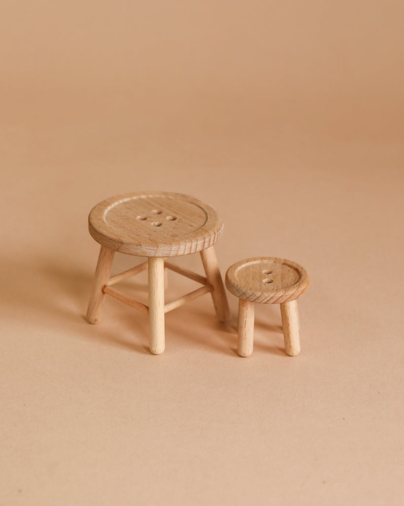 A photograph shows two small, round, wooden stools of different sizes on a plain beige background. The larger stool has four legs and a circular seat, while the smaller one—reminiscent of items found in the Maileg Bathroom Starter Set—has three legs and a similar round seat. Both seats feature subtle, carved details.