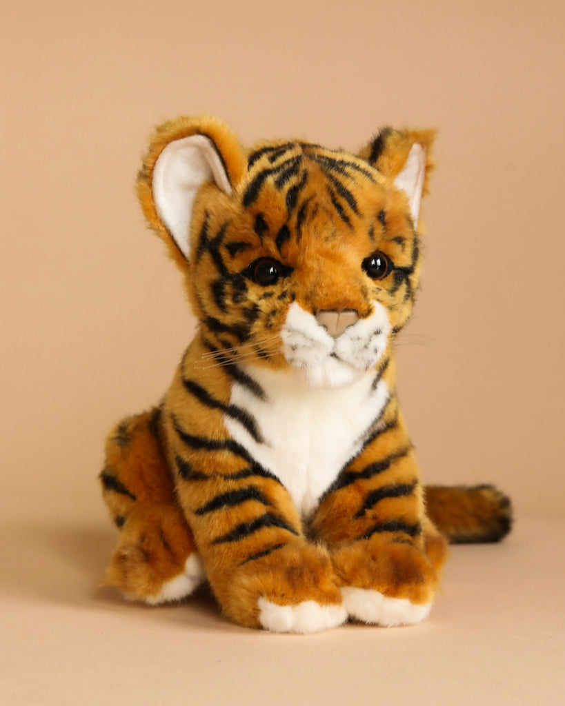 The Tiger Cub Stuffed Animal, with its realistic orange fur, black stripes, and white accents on its face and paws, sits on a neutral beige background. This hand-sewn plush toy features a cute, expressive face with black button eyes and a soft, fuzzy texture that gives it a unique personality.