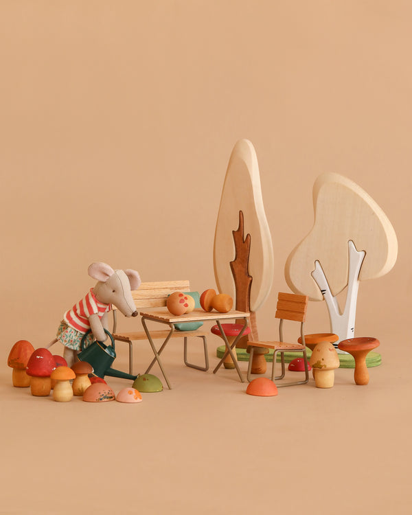 A Maileg Woodland Picnic Set features a plush Tricycle Mouse in a striped shirt and skirt watering Wooden Mushrooms by a miniature table and chairs, colorful mushrooms, and wooden trees, all arranged on a beige background.