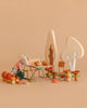 A Maileg Woodland Picnic Set features a plush Tricycle Mouse in a striped shirt and skirt watering Wooden Mushrooms by a miniature table and chairs, colorful mushrooms, and wooden trees, all arranged on a beige background.