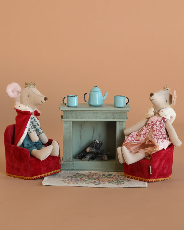 Maileg King and Queen Mouse wear crowns and royal outfits, seated in Maileg Soft Folding Chairs by a green fireplace. The Maileg Royal Tea Time Set with a blue teapot and cups rests on the mantle, atop a floral rug with a warm beige backdrop.