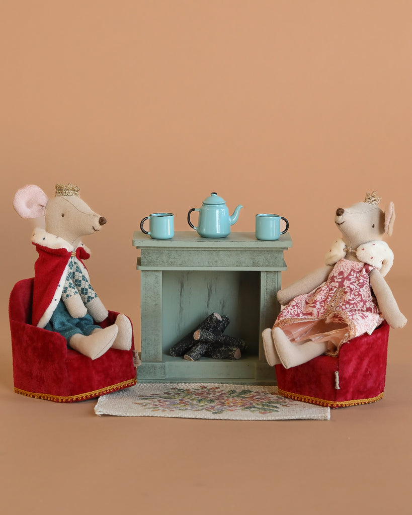 Maileg King and Queen Mouse wear crowns and royal outfits, seated in Maileg Soft Folding Chairs by a green fireplace. The Maileg Royal Tea Time Set with a blue teapot and cups rests on the mantle, atop a floral rug with a warm beige backdrop.