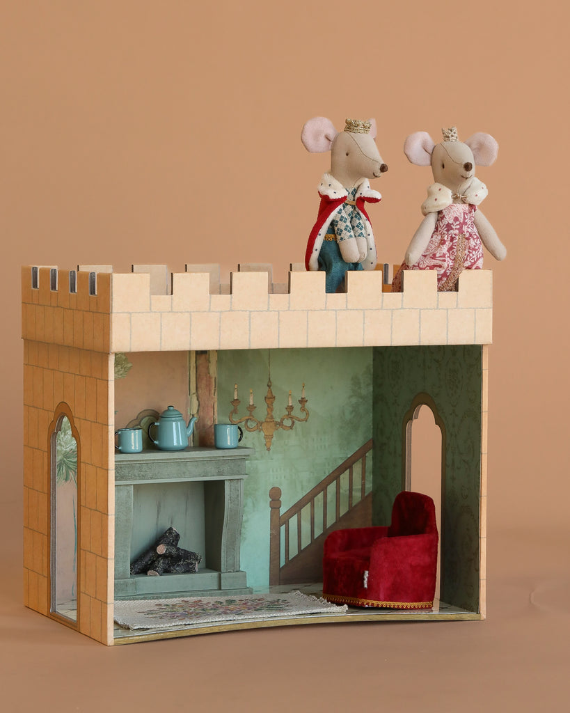 The Maileg King and Queen Castle Hall Set features royal mice with crowns and regal outfits atop a castle hall, complete with a fireplace, red armchair, and teapots inside. The backdrop is plain brown.