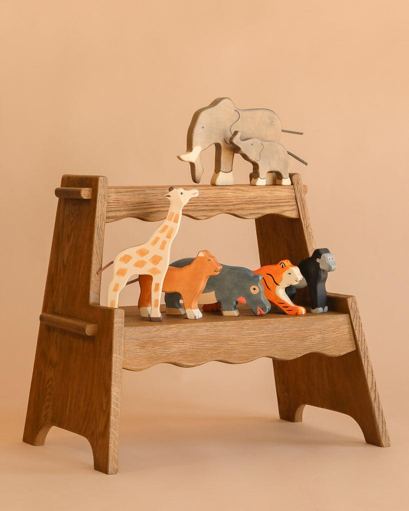 A wooden display shelf features the Holztiger Handmade Wooden Safari Animals Set—handcrafted elephant, giraffe, lion, hippo, tiger, and gorilla—made from maple and beech wood, set against a plain beige background.