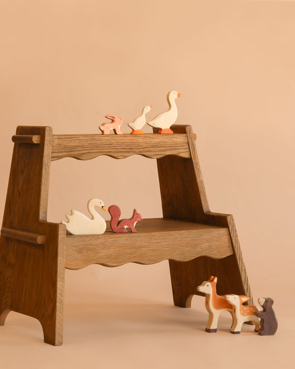 The Holztiger Handmade Wooden Woodland Animals Set features maple and beech wood animals—ducks, a swan, a squirrel, a rabbit, and two deer—displayed on a wooden two-tiered shelf against a beige background.