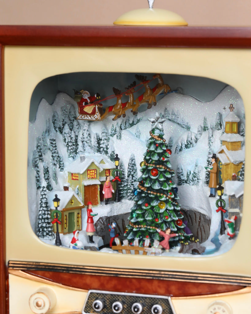 An animated Christmas Music TV With Skaters styled as a vintage television set, displaying a miniature Christmas village scene with a decorated tree, snow-covered houses, and Santa Claus in his sleigh flying above.