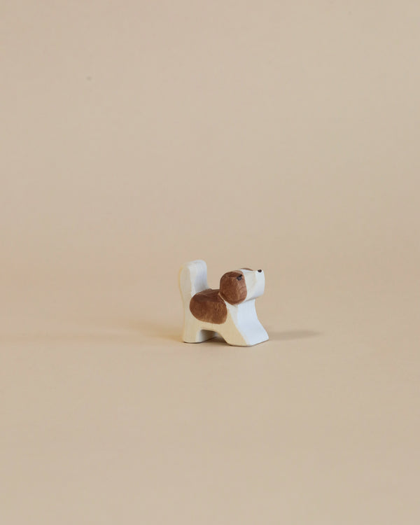 A small, handcrafted wooden figurine of a dog with patches of brown and white stands on a light beige background. The dog, an Ostheimer Small Saint Bernhard Dog - Head Up, has a simple, stylized design with a short tail and upright posture, perfect for imaginative play as it looks to the right.