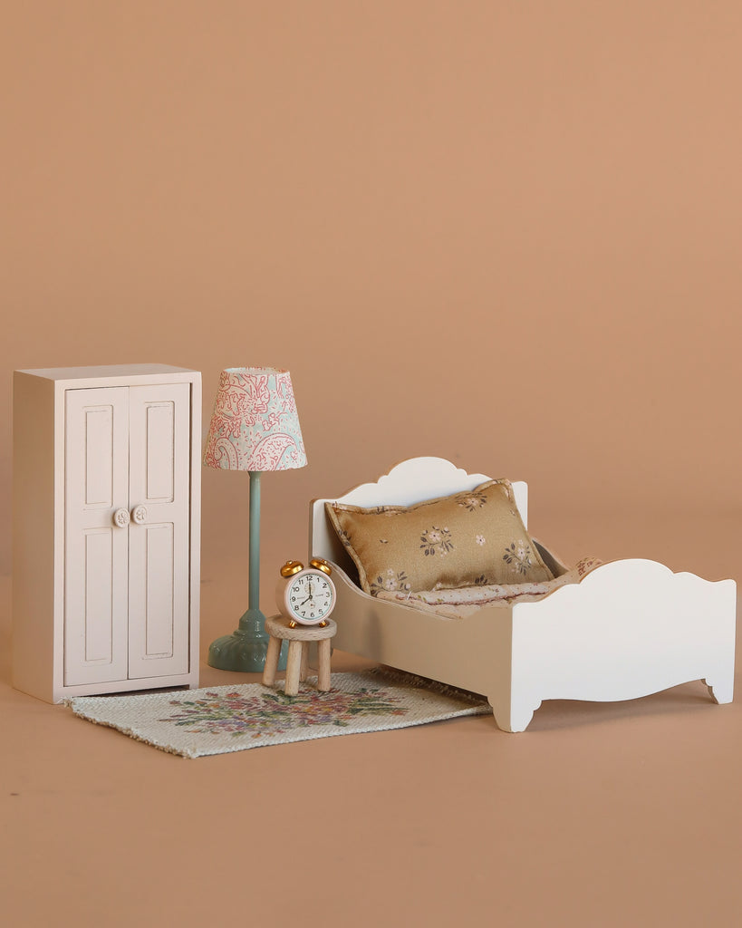 The Maileg Bedroom Set includes a wooden bed with a patterned pillow, a white wardrobe, a vintage floor lamp with pink shade, a tiny clock, and a floral rug, all displayed on a plain beige background.
