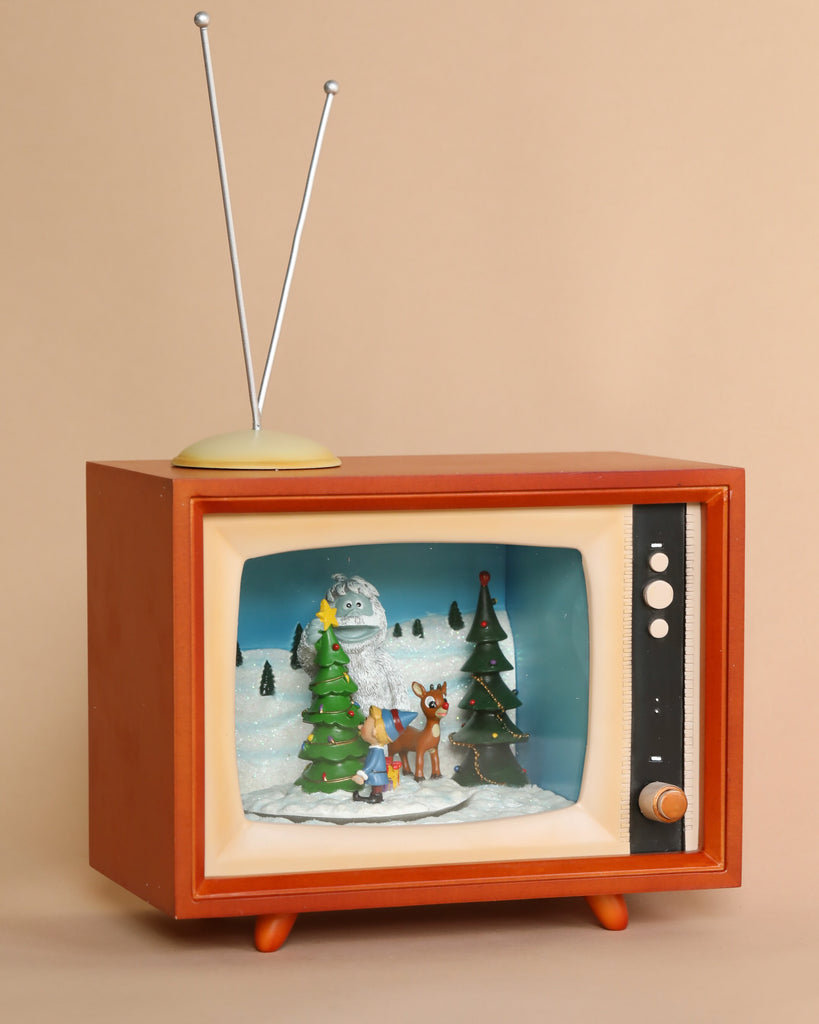 The Christmas Music TV With Rudolf features a vintage television set displaying a winter scene with a snowman, Christmas trees, and Rudolph The Red-Nosed Reindeer. An antenna is perched on top of the TV, and the beige background highlights the retro style of the illustration.