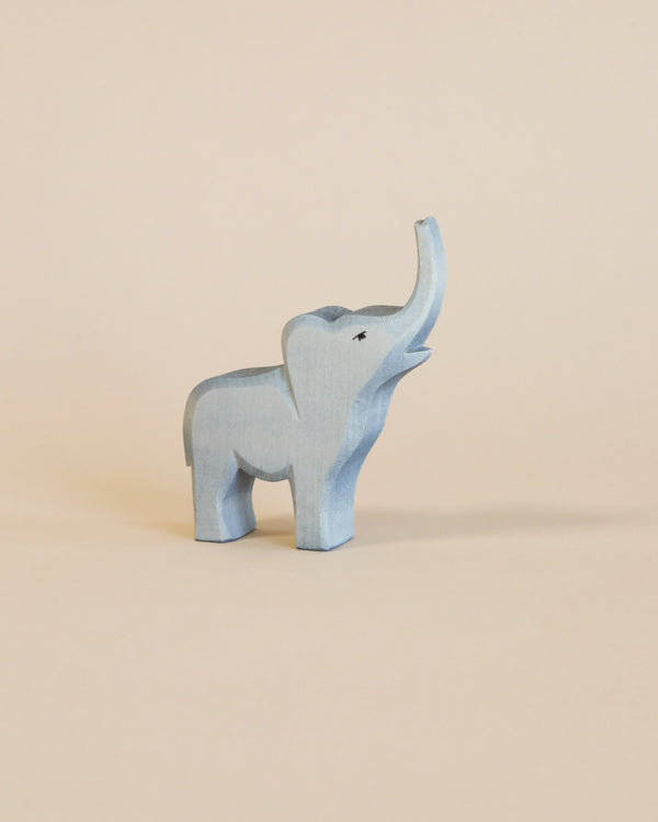 A small wooden Ostheimer Small Elephant - Trumpeting figurine stands against a plain, light beige background, showcasing detailed carvings and a soft blue-gray paint finish.