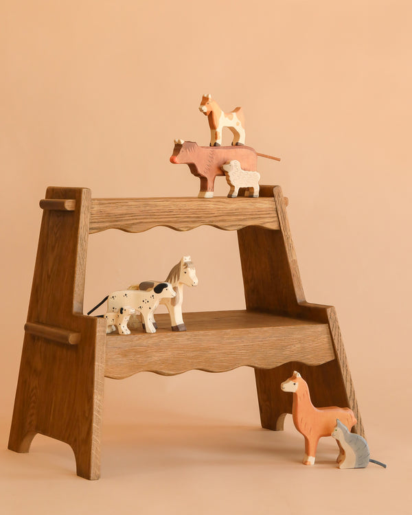 A wooden step shelf displays the Holztiger Handmade Wooden Farm Animals Set—handmade maple and beech wood figures like cow, sheep, cat, horse, dog, llama, and mouse—arranged on shelves against a beige background.