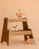 A wooden step shelf displays the Holztiger Handmade Wooden Farm Animals Set—handmade maple and beech wood figures like cow, sheep, cat, horse, dog, llama, and mouse—arranged on shelves against a beige background.