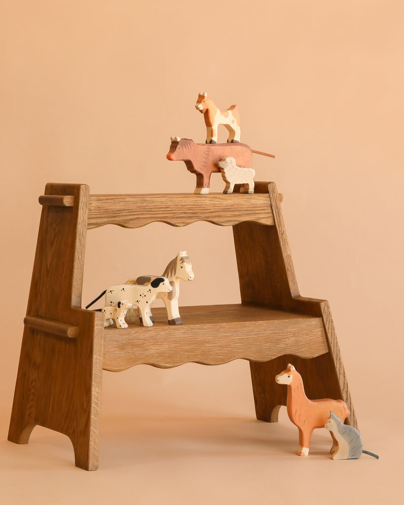 A wooden step shelf displays the Holztiger Handmade Wooden Farm Animals Set—handmade maple and beech wood figures like cow, sheep, cat, horse, dog, llama, and mouse—arranged on shelves against a beige background.