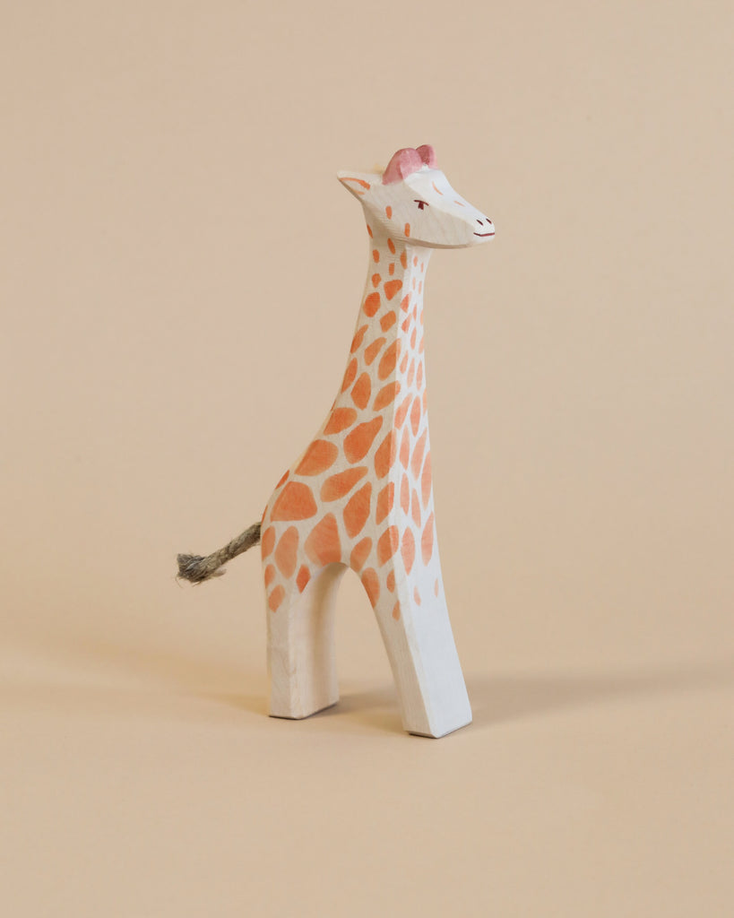 A charming, hand-painted Ostheimer Giraffe - Standing figurine with noticeable pink spots on its neck, a small hat, and a tiny tail, evoking the spirit of imaginative play, displayed against a pale