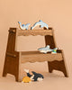 The Holztiger Handmade Wooden Sea Animals Set—featuring two dolphins, a shark, a whale, and a turtle—rests on a two-step wooden stool against a plain beige background. These toys are handcrafted from maple and beech wood.