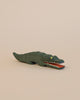 A small, handcrafted Ostheimer Crocodile with a simple design, painted green and featuring yellow eyes and red details inside its open mouth, set against a plain light background.