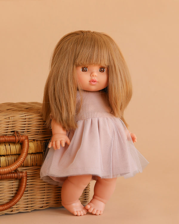 The Minikane Baby Doll (13") - Nina with Clothing has long, straight light brown hair with bangs and wears a pale pink dress. This anatomically correct doll stands barefoot by a wicker basket against a beige background.